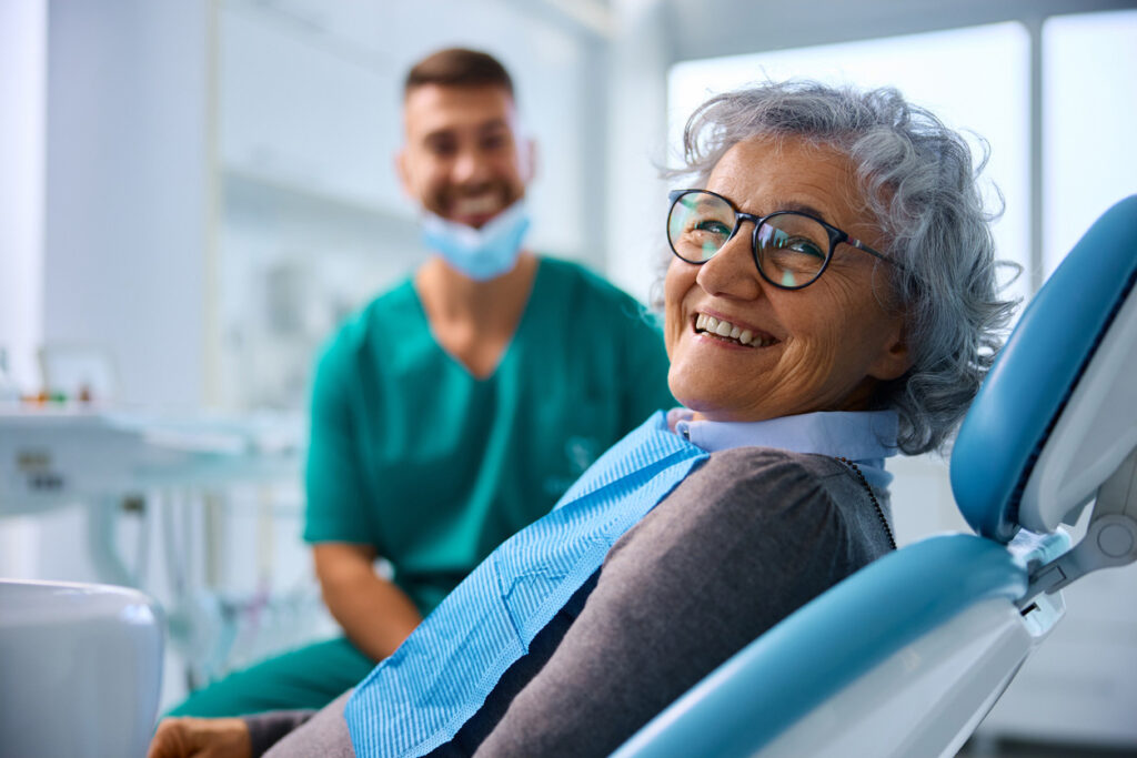 Patient sitting in dental chair looking across shoulder at camera. Dental professional in teal scrubs visible in background partially out of focus